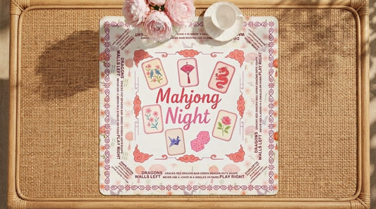Four women playing mahjong on a pink floral mat at a table inside a glass botanical conservatory