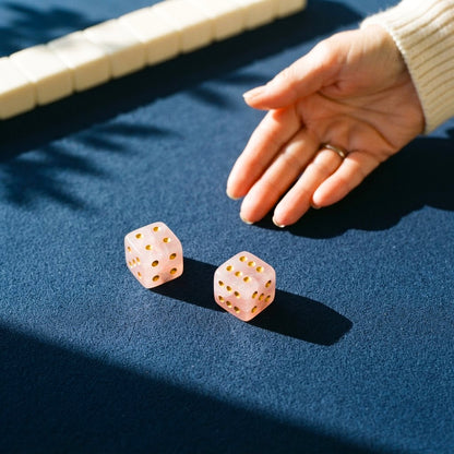 A hand beside two rose quartz mahjong dice just rolled onto a navy mat