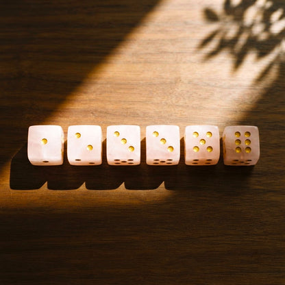 Set of six rose quartz mahjong dice with gold pips lined up on dark walnut