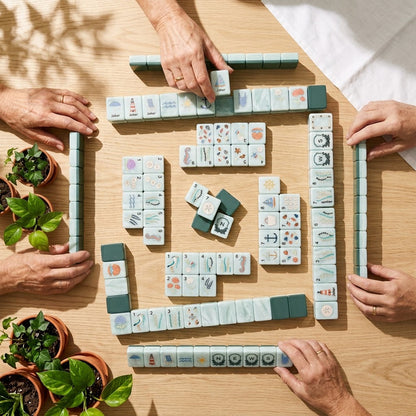 A Nantucket Shore game in play seen from above, four players' hands reaching in around the tiles