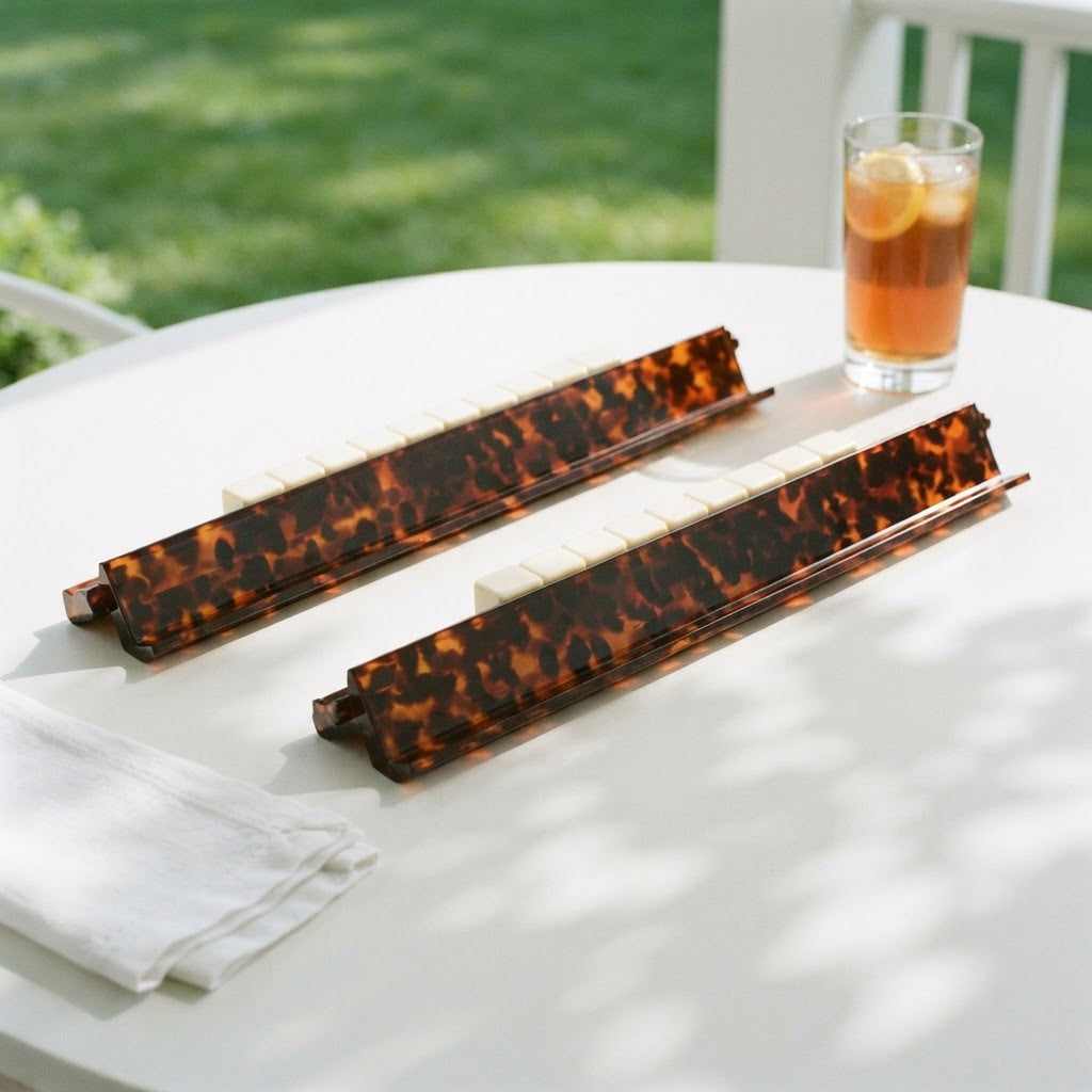 Two Tortoise Shell mahjong racks with tiles on a porch table beside a glass of iced tea